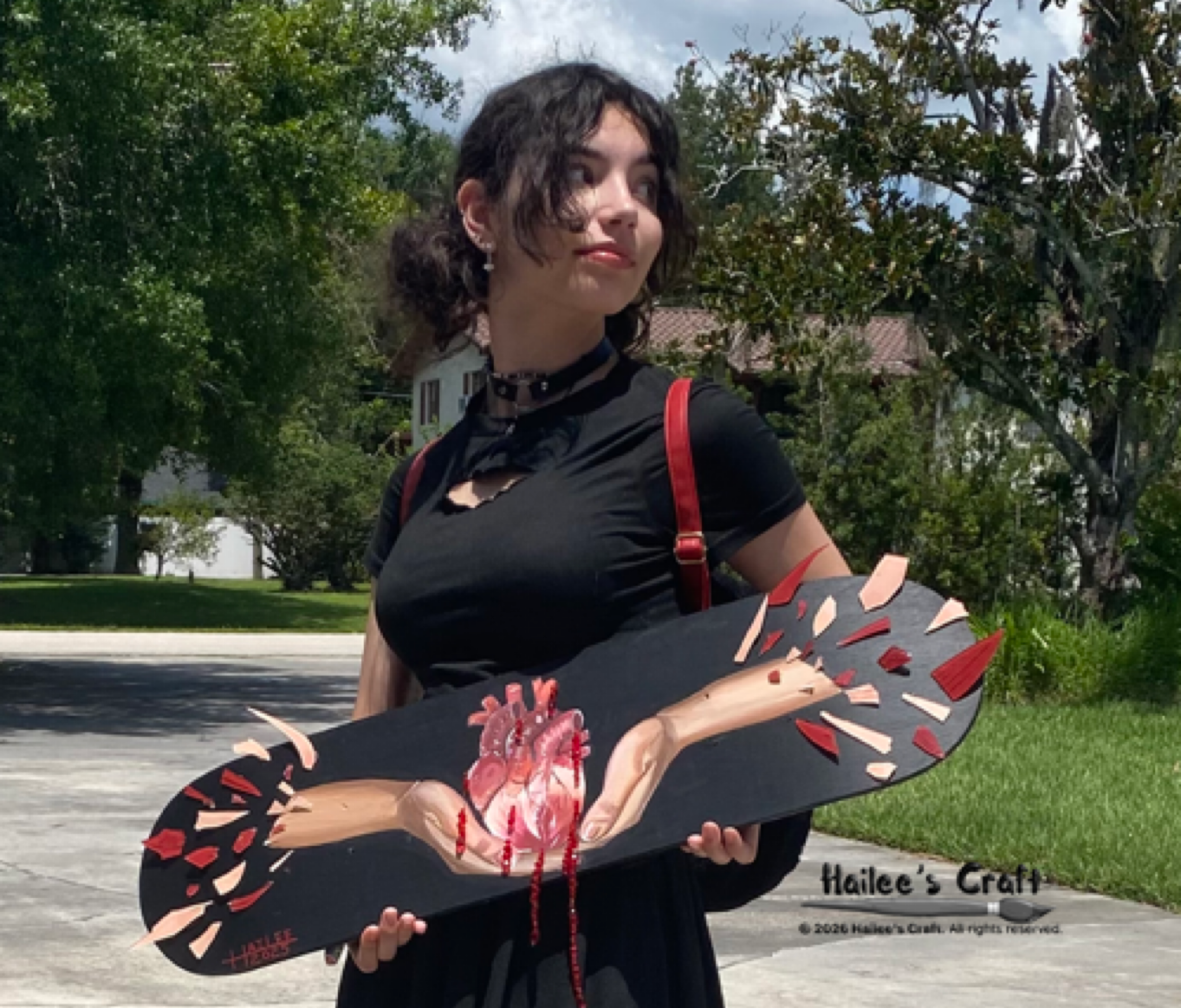 Hailee standing in a grassy area holding a skateboard. the skateboard is painted black, an in the center of the skateboard are two hands holding an atomically correct heart. shards of red and tan glass are sticking out of the arms of the painting. From the bottom of the heart, glass beads representing blood, are hanging down. 